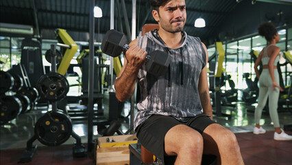 Asian athlete man in sportswear doing seated bicep arm curl with single dumbbell in fitness gym. Muscular male bodybuilder with serious face is lifting dumbbell to improve strength of bicep muscles.