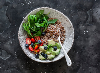 Fototapeta na wymiar Healthy balanced vegetarian breakfast - buckwheat with avocado, arugula, cherry tomatoes and blueberries on a dark background, top view