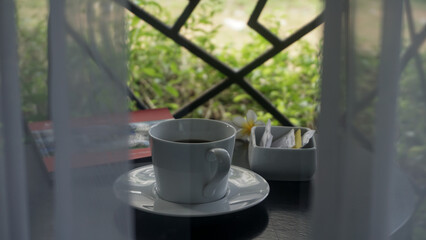 A cup of coffee served on the table at the hotel balcony captured from inside the room behind the window and white curtain