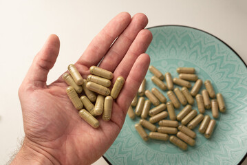 one hand holds some kratom pills with green plate with other kratom pills on background, selective focus