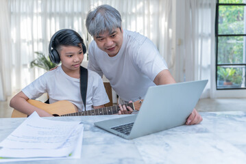 Asian teenage boy studying music, playing guitar online at home, with a father to take care of and help in learning, to Asian family and education online concept.