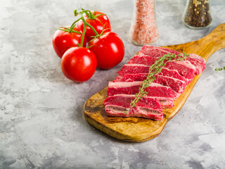 Appetizing raw meat steaks on a cutting board, ripe red tomatoes, spices in a glass jar on a gray background. The process of cooking homemade meat steaks. There are no people in the photo.