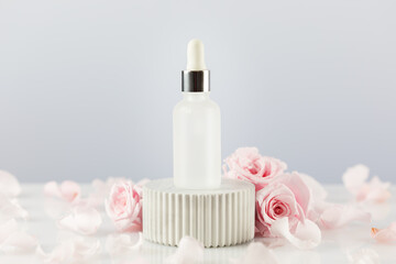 Fototapeta na wymiar Glass dropper bottle with serum on a marble podium on a white table with tender pink roses and rose petals. Natural beauty product based on rose flowers, fermented cosmetic. Soft focus style