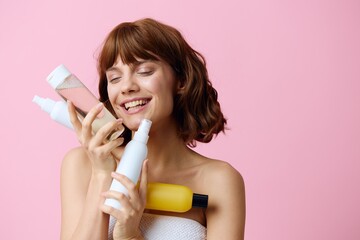  photo of a sweet, joyful woman standing on a pink background with different skin care products, smiling broadly at the camera, pressing them to the cheek