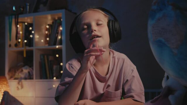 Little cute girl with headphones listen music and sing using a triangle ruler as a microphone at night. Schoolgirl doing homework and having fun