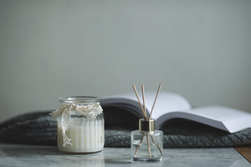 Glass bottle with scented candle and liquid home perfume over open paper book on table in room close up. Cozy hygge atmosphere.