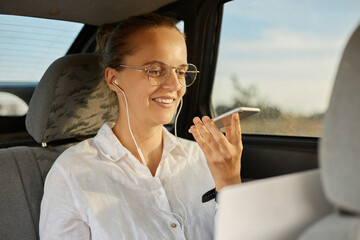 Portrait of smiling businesswoman sitting on back seat of car and working with laptop and holding...
