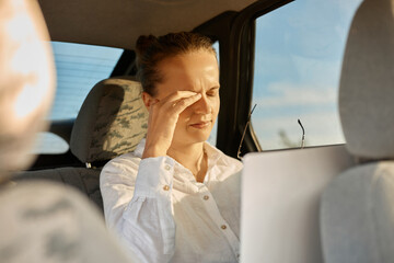 Portrait of exhausted business woman working laptop computer in car, female in white shirt using notebook in vehicle, typing on laptop at remote workplace, fells eyes hurt, rubbing her eye.