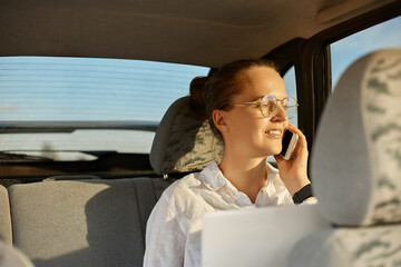 Portrait of attractive young adult woman in optical glasses sitting in the car and talking on the cell phone, holding laptop, looking away at the automobile window, enjoying sunset on her way home.