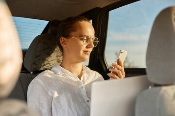Horizontal shot of woman in white shirt working laptop computer on back seat of car, using notebook and mobile phone in vehicle during her way to office or home.