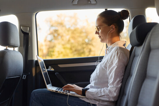 Side View Portrait Of Slim Businesswoman Wearing White Shirt Sitting On Back Seat Of Car And Working With Laptop, Listening Music Or Podcast On Her Way To Office, Sitting With Earphones.