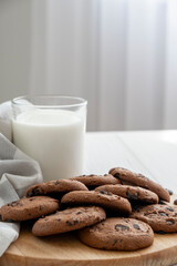 Morning breakfast of chocolate chip cookies on wooden board and glass of milk with tulle window on background. Recipe of wheat cookies with chocolate chips.