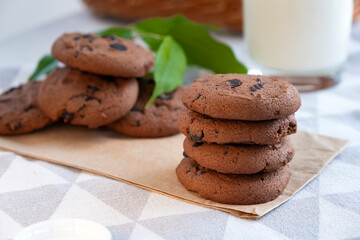Stack of chocolate chip cookies with glass of milk, biscuits and wicker basket on background. Recipe of delicious dessert for tea, freshly baked cookies with chocolate crumbs.