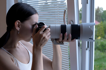 Young woman Paparazzi Photographer Capturing A Photo from an apartment window. Suspiciously Jealous wife Detective Spy Using A Big Professional Camera.