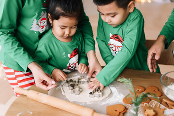 An Asian family with two children in green pajamas cook together festive cookies celebrate the Christmas holiday in decorated house in winter. Mom dad son and daughter spend time together on vacation 