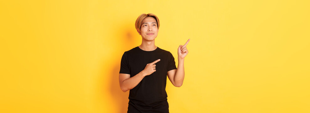 Thoughtful and pleased handsome asian man smiling and pointing fingers upper left corner, standing over yellow background