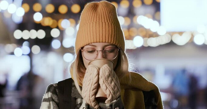 A young girl in glasses drinks a hot drink from a large paper cup, standing on the street in winter against the backdrop of city lights.