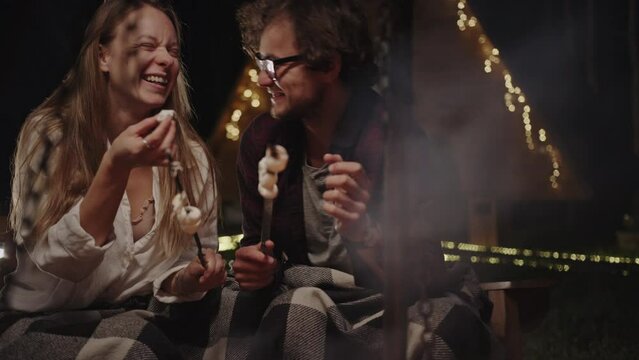 Cheerful romantic couple eating BBQ marshmallow near bonfire at night