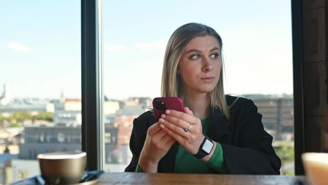 A beautiful, confident woman with a phone in her hands looks out the window against the background of the city panorama. Successful business woman, 
social media marketing manager