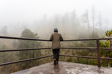 Fototapeta premium Tourist woman in brown coat is looking at the misty forest. Wanderlust travel concept, space for text, atmospheric epic moment, Nevado de Colima