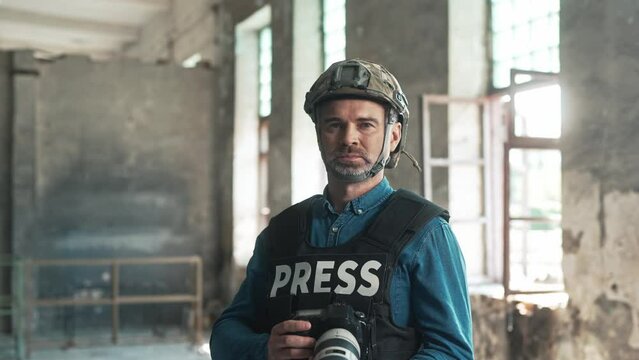Portrait of male war journalist wearing bulletproof vest and casque holing in hands photo camera taking pictures in battle area. Middle-aged professional man war press working in conflict area