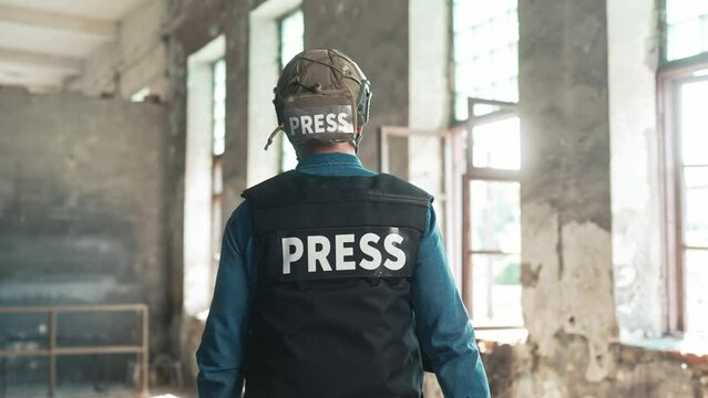 Back view of male war press correspondent with photo camera. Rear of man journalist in bulletproof vest and helmet in damaged building. Cameraman standing in battle area. Military news