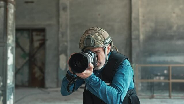 One man in helmet and bulletproof vest holding camera taking photos in war zone. Male war journalist with digital camera at the place of action in damaged building working. War press. Armed conflict