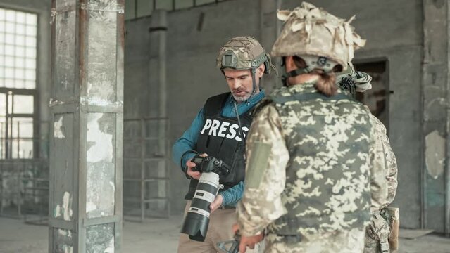 Portrait of middle-aged professional photographer journalist working at war zone taking photos of two brave soldiers in camouflage uniform and helmets in old ruined building. War press Photo reportage