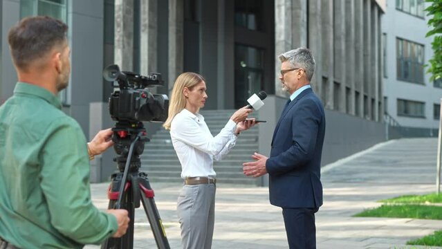 Caucasian beautiful woman TV correspondent holding smartphone and microphone interviewing middle-aged male politician standing in street. Handsome businessman talking on camera with female journalist