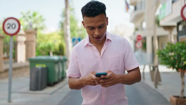 Young latin man smiling confident using smartphone walking at street