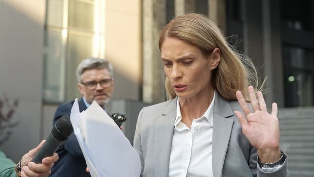 Close up of beautiful Caucasian female politician holding papers in hands in bad mood walking on street passing by journalists with microphones. No comments. Businesswoman doesn't want to comment