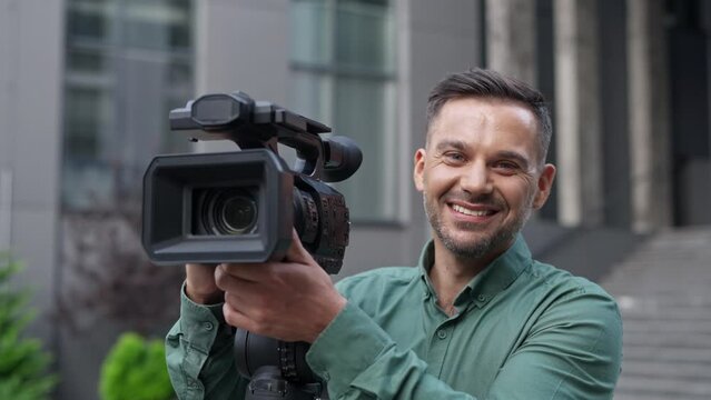 Close up of cheerful handsome Caucasian cameraman working outdoor in city using professional equipment. Happy young video operator shooting reportage smiling at camera. Broadcasting live in street