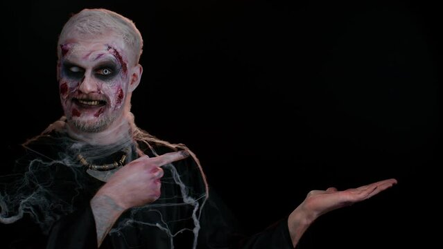 Creepy man with bloody scars face, Halloween stylish zombie make-up. Scary wounded undead guy showing thumbs up and pointing at right on blank space. Place for advertisement logo. Sinister monster