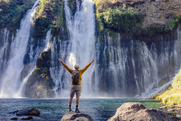 Man traveler standing with his hands raised with a view on the beautiful waterfall, view from the back