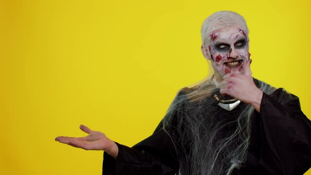 Creepy man with bloody scars face, Halloween stylish zombie make-up. Scary wounded undead guy showing thumbs up and pointing at left on blank space. Place for your advertisement logo. Sinister monster