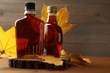 Bottles of tasty maple syrup and dry leaves on wooden table, space for text