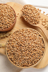 Wheat grains in bowls and spoon on wooden board, flat lay