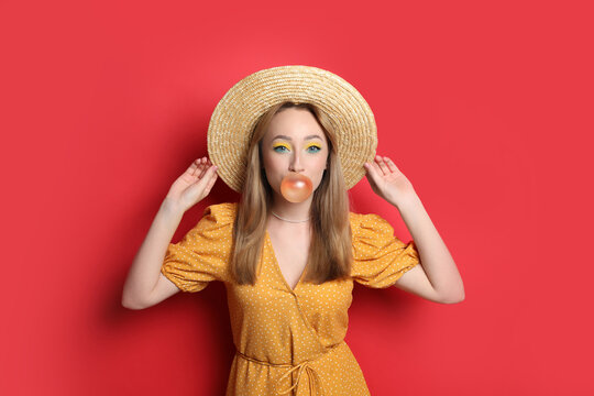 Fashionable young woman with bright makeup blowing bubblegum on red background