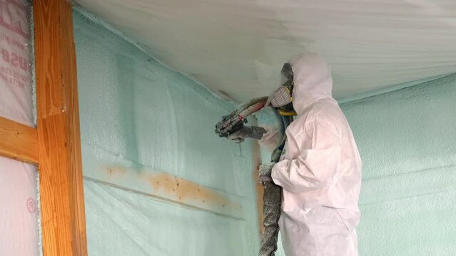 Worker in respirator and Tyvek suit sprays closed cell foam insulation near the ceiling of a newly constructed exterior wall; concepts of hazards, protection and new construction