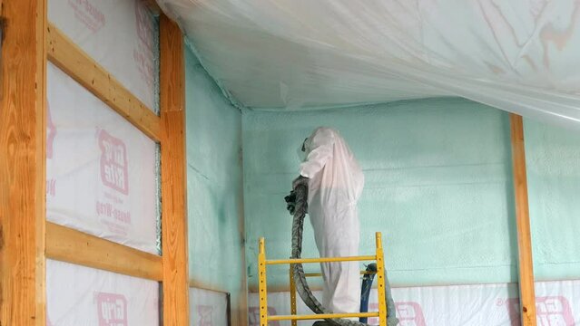 Worker in respirator and Tyvek suit sprays closed cell foam insulation on newly constructed exterior wall while standing on scaffolding; concepts of hazards, protection and new construction