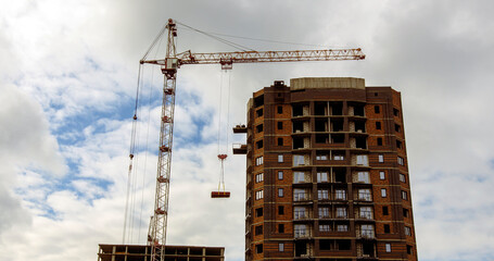 Fototapeta na wymiar High crane against cloudy sky. The low angle of inclination of a modern high crane and a multi-storey building under construction, located against a cloudy sky on the street