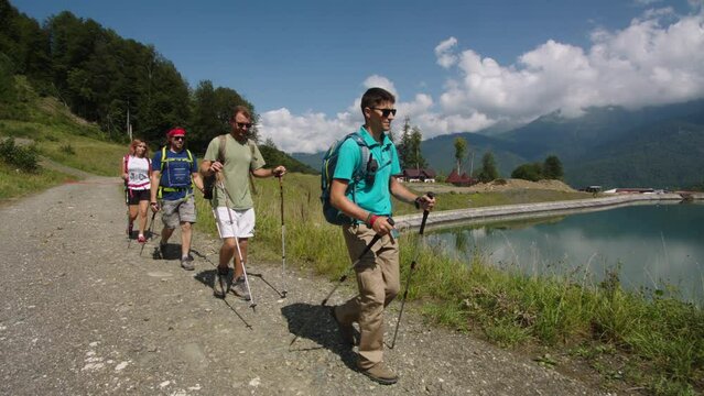 Group of young hikers walks down past transparent blue mountain lake. Instructor confidently leads group to goal. The fight against hypodynamia for white collars