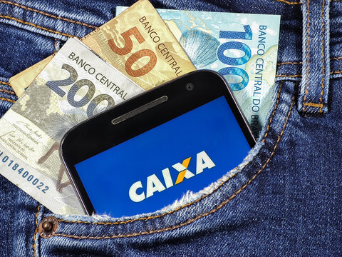 Smartphone with Caixa Econ&ocirc;mica Federal Bank logo on the screen in jeans pocket with notes of 100 reais 200 reais and 50 reais.