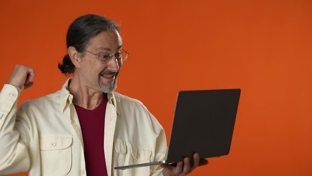Happy surprised mature bearded man using laptop say ye omg isolated on solid orange background studio portrait