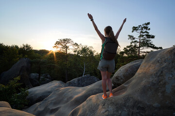 Sportive woman standing alone on hillside trail with raised up arms at sunset. Female hiker enjoying view of evening nature from rocky cliff on wilderness path. Active lifestyle concept