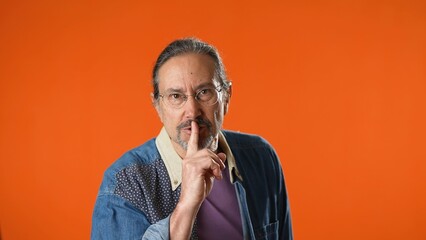 Secret quiet elderly gray-haired bearded man 50s 60s say hush be quiet with finger on lips shhh gesture isolated on plain solid orange background studio portrait