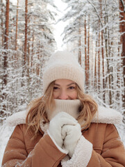 Winter walk, a young beautiful blonde in winter clothes walking in a snowy forest, a beautiful frosty day. A young woman warms her nose wrapped in a white scarf