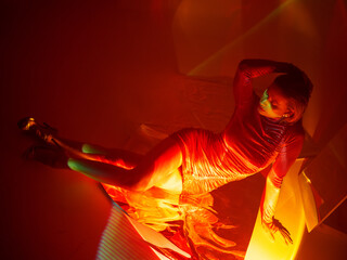 Glamorous portrait of a young beautiful blonde in a stylish scarlet short dress, sitting on the floor, refraction effects of colored red light, futuristic fashion shooting in neon light
