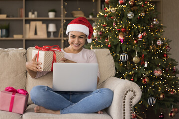 Happy Indian Xmas girl making video call from home, using laptop computer on couch at Christmas tree, holding gift box, thanking for present. Young woman, blogger recording festive New Year post