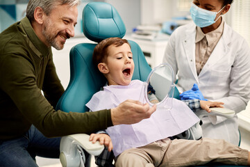 Small boy looking his teeth in mirror while being with his father at dentist's.